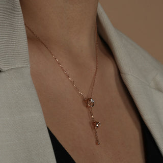 A close-up photo of a woman's neck wearing a grey suit wearing a delicate rose gold necklace adorned with two interconnected circles, each featuring a sparkling diamond in the centre. The image features the close-up front view of a swirling 14 KT Diamond Necklace, adorned with 0.21 carats of lab-grown diamonds, elegantly worn on the neck of the model of size 16 secured through a spring lock weighing a total of 3.28 grams. 