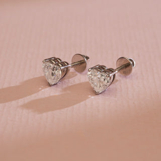 Cupid’s Heart Diamond Stud Earrings on pink background, crafted in 14KT hallmarked white gold. Features EF VVS solitaire diamonds totaling 0.70 carats, including a 0.35-carat stone. Bombay screw closure ensures secure wear. Lightweight at 1.21 grams, priced INR 15,000–50,000.