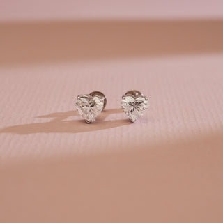 Front view of Cupid’s Heart Diamond Stud Earrings photographed on a soft pink background. 14KT white gold with EF colour, VVS clarity diamonds totaling 0.70 carats, solitaire 0.35 carats. Bombay screw closure, 1.21 grams, price INR 15,000–50,000.