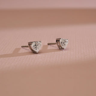Side angle of Cupid’s Heart Diamond Stud Earrings against pastel pink background. Crafted in 14KT hallmarked white gold, set with EF VVS diamonds weighing 0.70 carats total, solitaire 0.35 carats. Product weight 1.21 grams, priced INR 15,000–50,000.