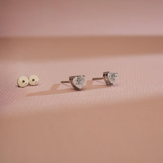 Cupid’s Heart Diamond Stud Earrings displayed on pink backdrop with visible 18KT hallmarked backs. Designed in white gold, featuring EF VVS clarity diamonds of 0.70 carats total, solitaire 0.35 carats. Secured with Bombay screw closure, weighs 1.21 grams, priced INR 15,000–50,000.