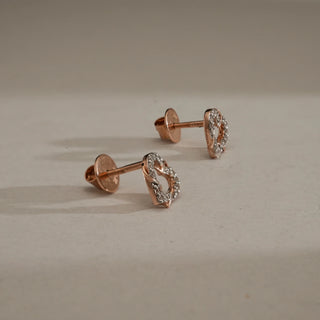Side view of Interlocked Heart diamond stud earrings in 14KT Rose Gold, placed on a neutral surface, featuring entwined heart motifs adorned with EF-VVS lab-grown diamonds and screw-back posts.