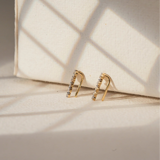 Propped against a softly lit cream surface, a pair of 14 KT yellow gold ear climbers earrings is arranged in a clean vertical stance. Each earring showcases a linear ascent of EF-color, VVS-clarity round diamonds totalling 0.310 carats, set along a sleek gold spine. The minimalist silhouette is elevated by precise stone alignment, creating a modern yet delicate statement. With a gross weight of 0.678 grams and a net weight of 0.616 grams, these earrings offer lightweight elegance and a subtle look.