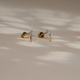 Displayed on a softly dappled beige surface, a pair of 14 KT rose gold diamond stud earrings is shown in a balanced front view. Designed in a nature-inspired silhouette, each earring features a graceful gold stem tipped with two marquise-shaped leaf motifs, encrusted with EF-color, VVS-clarity round diamonds totalling 0.060 carats. The minimal and organic design offers a delicate sparkle with modern charm. Weighing 0.66 grams gross and 0.648 grams net, these earrings are perfect for everyday wear. 