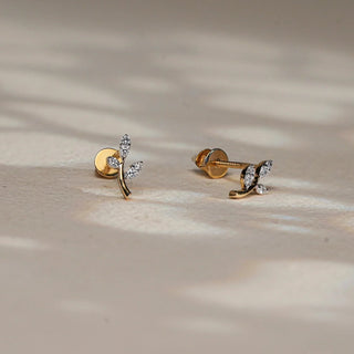 Resting on a softly illuminated beige surface, a pair of 14 KT rose gold diamond stud earrings is shown in angled and side views. Each earring is designed as a delicate gold stem branching into two marquise-shaped diamond-studded leaves, crafted with EF-color, VVS-clarity round diamonds totalling 0.060 carats. The screw-back posts ensure comfort and security, while the organic silhouette lends a touch of whimsy and refinement. With a gross weight of 0.66 grams and net weight of 0.648 grams. 