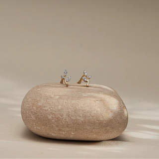 Displayed atop a smooth, neutral-toned stone against a soft beige background, a pair of 14 KT rose gold diamond stud earrings appears in a perfectly aligned front-facing view. Each earring features a curved gold stem tipped with two marquise-shaped diamond leaves, crafted using EF-color, VVS-clarity round diamonds totaling 0.060 carats. The delicate, botanical design adds a natural elegance to the lightweight structure. With a gross weight of 0.66 grams and net weight of 0.648 grams. 