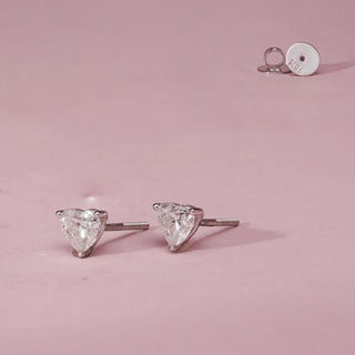 Side-angle view of Reiaâ€™s Heart-Cut Minimalist Diamond Earrings placed on a pink surface, highlighting the sleek white gold prong setting, heart-shaped diamond profile, and secure Bombay screw fastenings