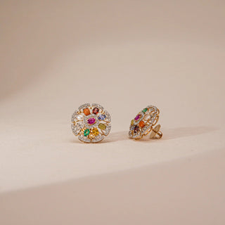 Angled front-side view of Reia's Nine-Gem Halo Heritage Earrings on a beige surface, highlighting the dimensional arrangement of colorful Navratna gems within a shimmering marquise-diamond halo. The tri-gold metal glows softly, symbolizing the fusion of cosmic symbolism and modern luxury.
