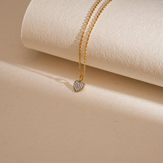 Gold chain necklace with a heart-shaped cluster pendant composed of small round diamonds, displayed against a soft ivory backdrop.