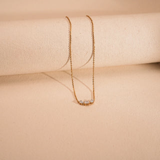 An elegant 14 KT yellow gold necklace, with a gross weight of 2.176 grams, is displayed stretched out and centrally against a smooth, light beige background. The delicate chain leads to a rectangular bar pendant, expertly set with sparkling emerald-cut VVS EF diamonds, totaling 0.940 carats, arranged in a continuous line.