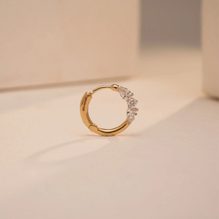 A fine Reia nose pin made of 18KT yellow gold, featuring three pear-shaped lab grown diamonds with an EF color and VVS clarity totaling 0.140 carats. The hoop stands upright on a light beige background basked in golden light—showcasing the beautiful curve of the hoop, and twinkling cluster of diamonds.