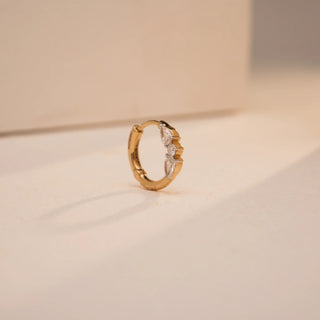 Reia's 18KT yellow gold push and pull nose pin showcases a set of three pear-shaped lab-grown diamonds (0.140 carats) set with an asymmetric orientation. Pictured from the side, on a sunlit cream background, the diamonds glimmer with the sunlight across the curved golden hoop.