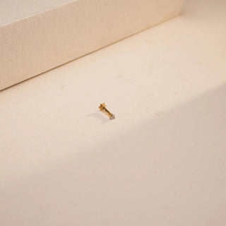 Wide-angle top view of the Lustre Single-stone Nosepin by Réia, placed on a softly lit cream-colored textured surface with a raised background element. Crafted in 18 KT yellow gold, the nosepin features a single round lab-grown diamond set in a minimalist frame with a South screw-back. The 0.04-carat diamond (EF color, VVS clarity) subtly sparkles, highlighting the piece’s elegant simplicity and refined craftsmanship.