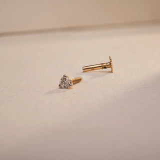 An image of Réia's 14 KT yellow gold nosepin, taken from above with a close-up, in dim light on a cream background. The pin has a single round, lab-grown diamond in a four-prong setting with a separate flat-back post. The diamond sparkles softly, off of E-F colors and VVS clarity. The flowers and the lighting give life to the nuanced clean shape and simplicity of this elegant, everyday design. 