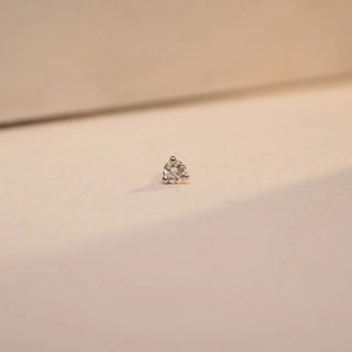 : A close-up view of a 14 KT yellow gold nosepin by Réia, featuring a single round lab-grown diamond with EF color and VVS clarity, secured in a four-prong setting. The nosepin is displayed on a softly lit, cream-toned surface, alongside a separate flat-back post, emphasizing its simple elegance and subtle sparkle