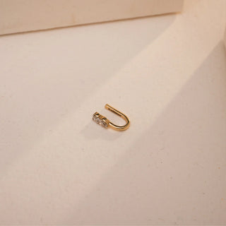 Reia’s 14KT yellow gold hanging nose pin, set with 0.070 carats of EF color, VVS clarity lab grown diamonds, rests elegantly on a neutral-toned flat surface. The pear-cut and round diamonds shimmer softly under warm, directional light in this graceful U-shaped silhouette