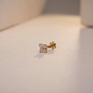 Close-up image of the Petite Blossom Gold Nosepin by Réia, crafted in 14 KT yellow gold and displayed on a softly lit, textured surface. The nosepin features a delicate four-petal floral design made from round lab-grown diamonds, totaling 0.08 carats (EF color, VVS clarity). The piece includes a South screw-back for secure wear and exudes subtle elegance and timeless femininity.