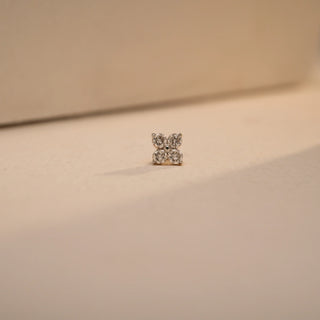 Front-facing close-up of the Petite Blossom Gold Nosepin by Réia, featuring a delicate four-petal floral design made from sparkling round lab-grown diamonds. Displayed on a softly lit cream-toned surface with gentle shadows, the nosepin is crafted in 14 KT yellow gold and showcases a total diamond weight of 0.08 carats (EF color, VVS clarity), exuding elegance and feminine charm.