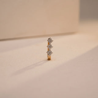 A close-up view of a delicate 18 KT yellow gold nose pin, weighing 0.47g, showcasing its prominent 0.060 carat luminous round EF VVS diamond. The nose pin is positioned on a light beige textured fabric, creating soft shadows, with a blurred light background, highlighting its detailed craftsmanship and continuous brilliance.