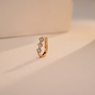 A tiny, dazzling 18 KT yellow gold nose pin, gross weight 0.47g, designed with a prominent 0.060 carat round EF VVS diamond. The nose pin is presented on a textured dark surface, with a blurred background, drawing focus to its elegant design and singular diamond.