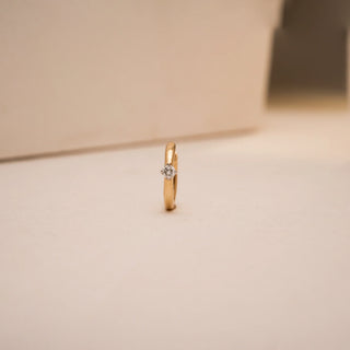 A minimalist 14KT yellow gold nose pin by Reia featuring a single round EF color, VVS clarity lab grown diamond of 0.04 carats. The sleek band stands upright on a soft beige backdrop, surrounded by warm, diffused light that enhances its polished elegance and everyday charm.