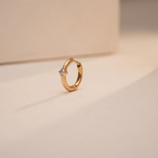 Reia’s 14KT yellow gold nose pin with a prong-set 0.04 carat round EF color, VVS clarity lab grown diamond, styled in a smooth hoop silhouette. Angled softly on a sunlit cream-toned base, the piece casts a gentle shadow, capturing both precision and subtle sparkle.
