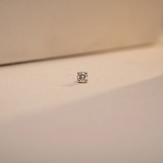 A minimalist Reia nose pin in 14KT yellow gold starring a brilliant round lab grown diamond solitaire of 0.070 carats (EF color, VVS clarity). Positioned on a soft beige surface and capturing it from the front, the image cultivates beauty in the clean prong setting of the diamond, its symmetry.