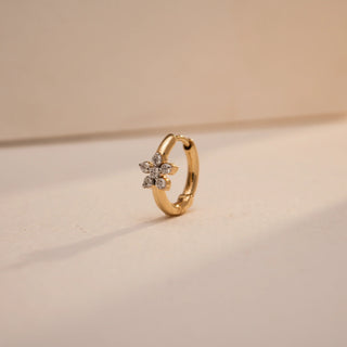 The nose pin is positioned at a diagonal angle, highlighting the gold hoop structure and the sparkle of the floral diamond cluster. The soft-focus background and warm lighting enhance the brilliance of the diamonds and the smooth gold finish.
