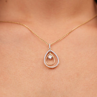 A close-up photo of a woman's neck wearing a delicate yellow gold necklace adorned with a teardrop-shaped pendant set with sparkling diamonds. The pendant features a double-layer design, with a smaller diamond in the centre surrounded by a larger diamond-encrusted oval. Close-up front view of this beautiful female necklace secured with a lobster lock crafted from 14 KT yellow gold and features 0.63 carats of diamonds of size 22