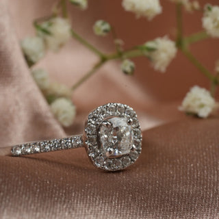 A close-up view of an ornate diamond engagement female ring of size 11 with a halo design, presented on a pinkish satin background. The centre stone is a radiant-cut diamond surrounded by a halo of smaller round brilliant diamonds set in 14 KT white gold. The total lab-grown diamond carat weight is 0.83 carats with a solitaire of 0.54 carats and the weight of the product is 1.72 grams, providing a brilliant and sparkling appearance to this elegant ring.