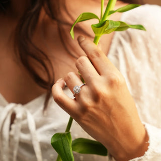 A view of an ornate diamond engagement female ring with a halo design modelled on a hand of size 11. The centre stone is a radiant-cut diamond surrounded by a halo of smaller round brilliant diamonds set in 14 KT white gold. The total lab-grown diamond carat weight is 0.83 carats with a solitaire of 0.54 carats and the weight of the product is 1.72 grams, providing a brilliant and sparkling appearance to this elegant ring.