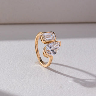 Side view of Toi Et Moi Diamond Ring in 14KT Yellow Gold, featuring an emerald-cut and pear-shaped lab grown diamond in bezel settings. Set on a beige backdrop, the design highlights depth and modern elegance.

