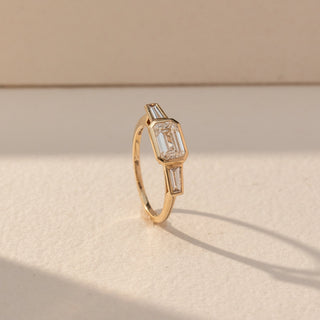 Side view of Emerald Cut Bezel Diamond Ring in 14KT Yellow Gold, featuring a centred emerald-cut lab grown diamond with tapered side stones. Set on a beige backdrop, the design highlights structure and refined elegance.
