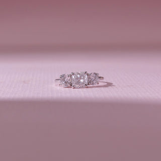 Front view of a three-stone diamond ring in white gold, featuring a radiant-cut centre diamond flanked by round diamonds. Captured on a soft pink backdrop, highlighting timeless brilliance.
