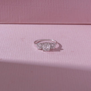 Top view of a three-stone diamond ring in white gold, featuring a radiant-cut centre diamond flanked by round diamonds. Captured on a soft pink backdrop, highlighting classic sparkle.