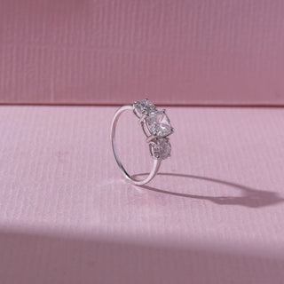 Angled view of a three-stone diamond ring in white gold, featuring a radiant-cut centre diamond with round side stones. Captured on a soft pink backdrop, highlighting refined brilliance and elegance.
