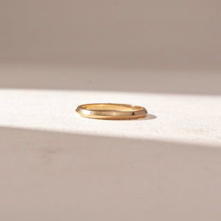 Front view of Minimal Faceted Band Ring in 14KT Yellow Gold, featuring a sleek geometric profile with polished facets. Set on a soft beige surface, the design reflects clean lines and understated shine.
