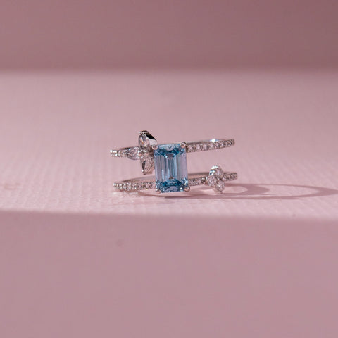 Front view of Emerald Cut Blue Stone Ring in 14KT White Gold, featuring a centred emerald-cut blue gemstone with lab grown diamond accents. Set on a pink backdrop, the design highlights contrast and elegance.
