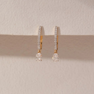 Front view of diamond hoop drop earrings in 14KT yellow gold, featuring pavé-set hoops with dangling round diamonds, displayed against a beige backdrop highlighting refined sparkle and elegance.
