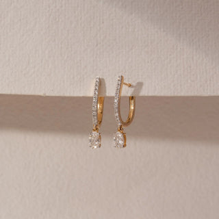 Angled view of diamond hoop drop earrings in 14KT yellow gold, featuring pavé-set hoops with dangling round diamonds, displayed against a beige backdrop highlighting depth and elegant sparkle.
