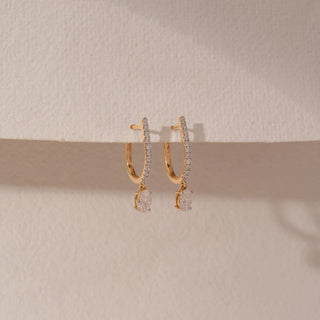 Side view of diamond hoop drop earrings in 14KT yellow gold, featuring pavé-set hoops with dangling round diamonds, displayed against a beige backdrop highlighting delicate brilliance and elegance.

