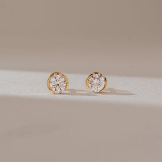 Front view of solitaire diamond stud earrings in yellow gold, featuring round lab-grown diamonds in a curved bezel-style setting. Captured against a soft beige background with gentle shadows.