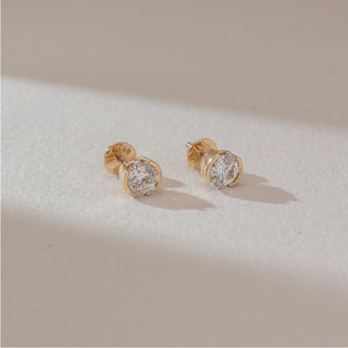 Angled view of solitaire diamond stud earrings in yellow gold, featuring round lab-grown diamonds in a curved bezel-style setting. Set against a soft beige textured background.