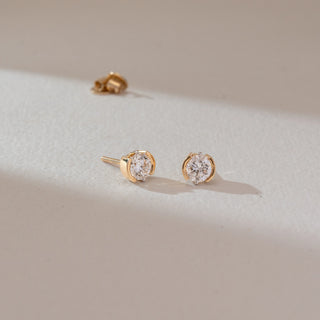 Side angled view of solitaire diamond stud earrings in yellow gold, featuring round lab-grown diamonds in a curved bezel-style setting. Captured against a soft beige textured background.