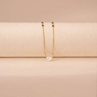 Front view of a 14KT yellow gold mangalsutra featuring a heart-cut diamond pendant with black beads, set against a beige background.
