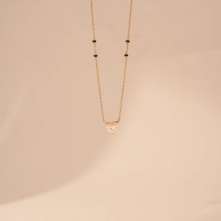 Front view of a 14KT yellow gold mangalsutra featuring a heart-cut diamond pendant with black beads, set against a beige background.
