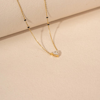 Angled view of a 14KT yellow gold mangalsutra featuring a heart-cut diamond pendant with black beads, set against a beige background.