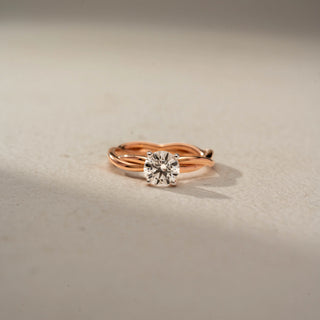 Front view of a round solitaire engagement ring in 14 KT Rose Gold, featuring a round lab-grown diamond set on an elegant twisted band, displayed against a beige backdrop.