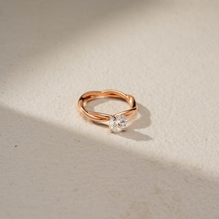 Side view of a round solitaire engagement ring in 14 KT Rose Gold, featuring a round lab-grown diamond set on an elegant twisted band, displayed against a beige backdrop.