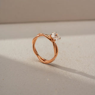 Angled view of a round solitaire engagement ring in 14 KT Rose Gold, featuring a round lab-grown diamond set on an elegant twisted band, displayed against a beige backdrop.